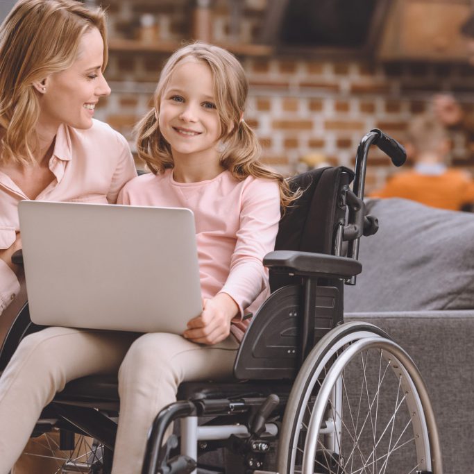 happy mother looking at cute little daughter sitting in wheelchair and using laptop at home