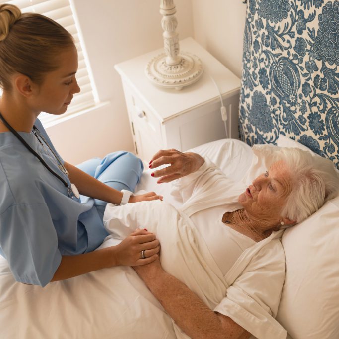 Side view of a senior woman lying while interacting with beautiful female doctor in bedroom at home